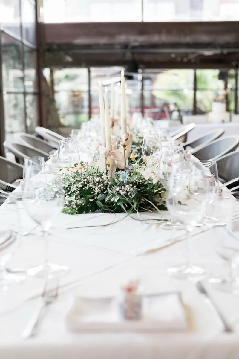 Long reception table set with a greenery runner, tall taper candles and glassware, windows onto the water behind it