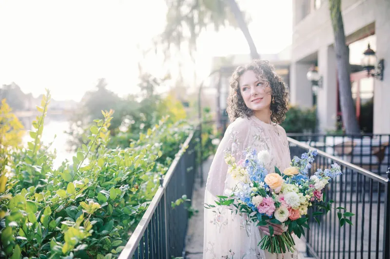 Bride standing at the iron railing on the lakeside walkway with her bouquet, late sun coming across the water behind her
