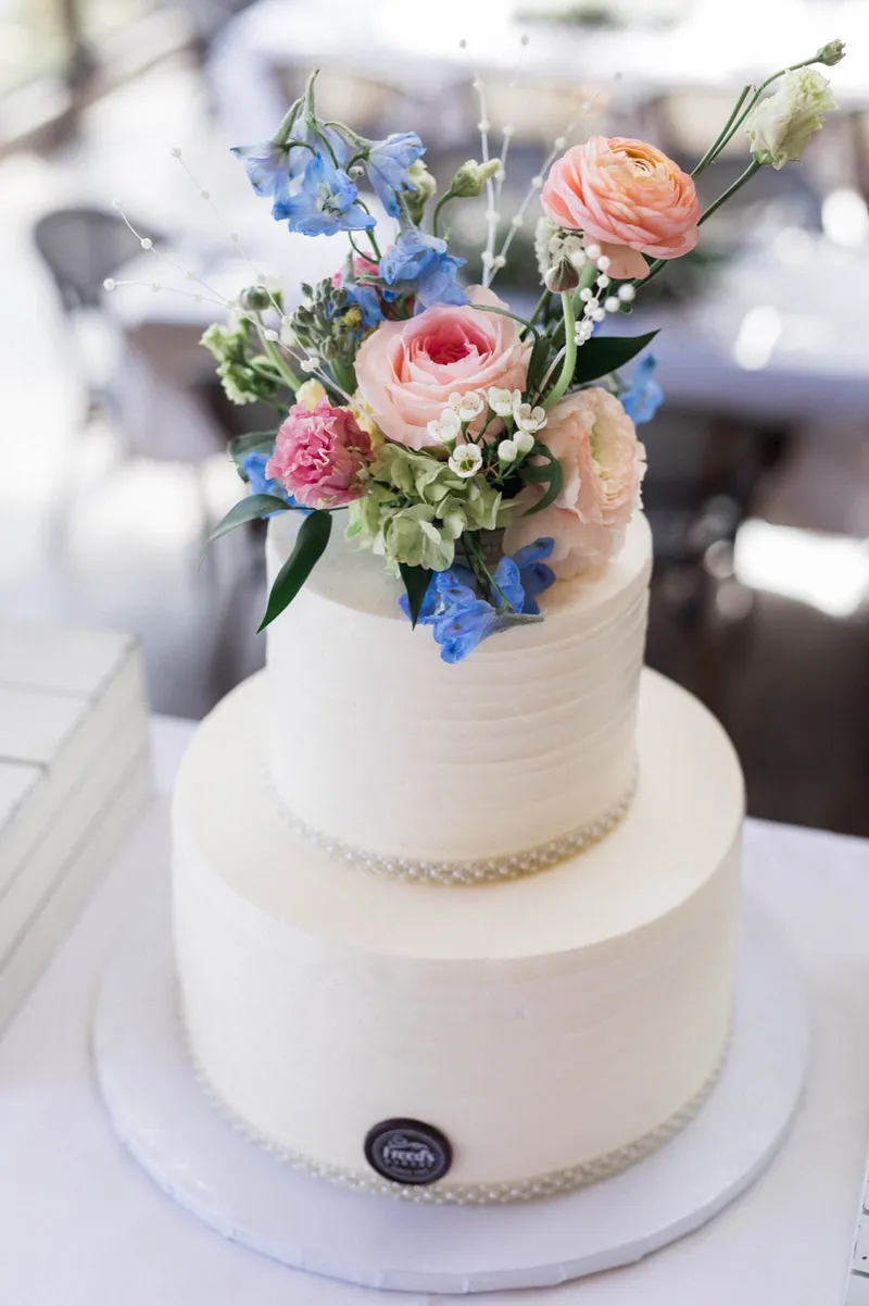 Two tier white cake topped with peach ranunculus, blue delphinium and pink roses