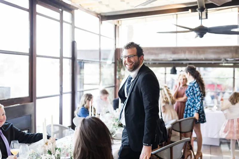 A guest in a dark suit turning to laugh at something across the reception, seated tables and glass doors behind him