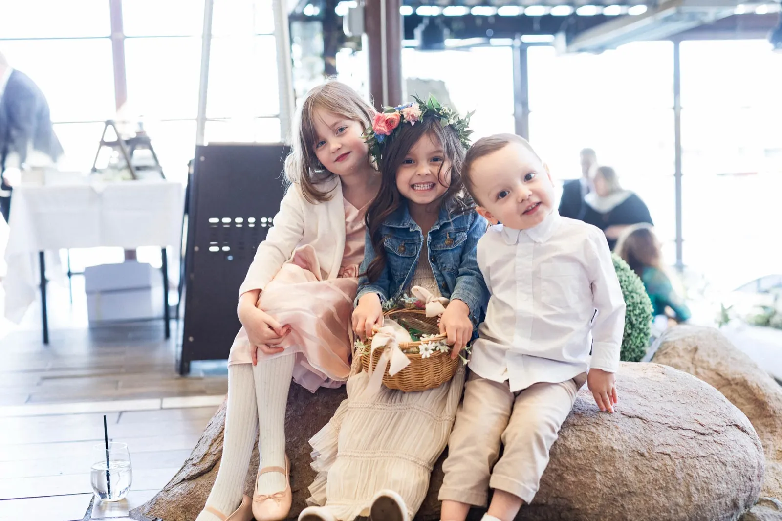 Three small children squeezed onto a boulder, one holding a basket of petals, all looking done with waiting