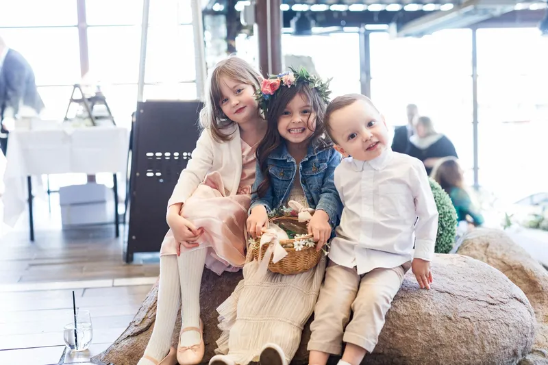 Three small children in wedding clothes sitting together on a low rock with a flower basket between them