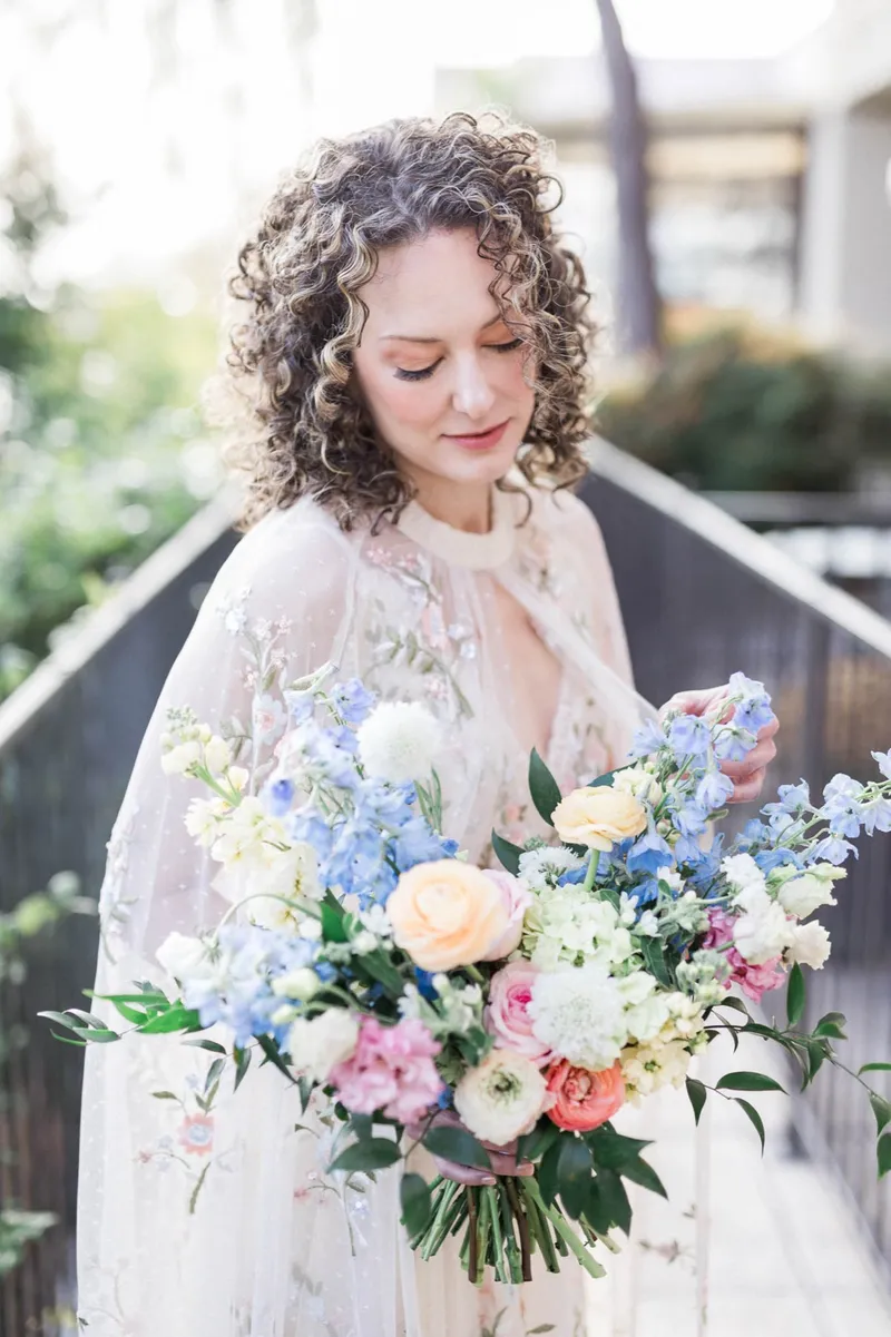 Bride looking down at her bouquet on the walkway, the cape falling off her shoulders in soft afternoon light
