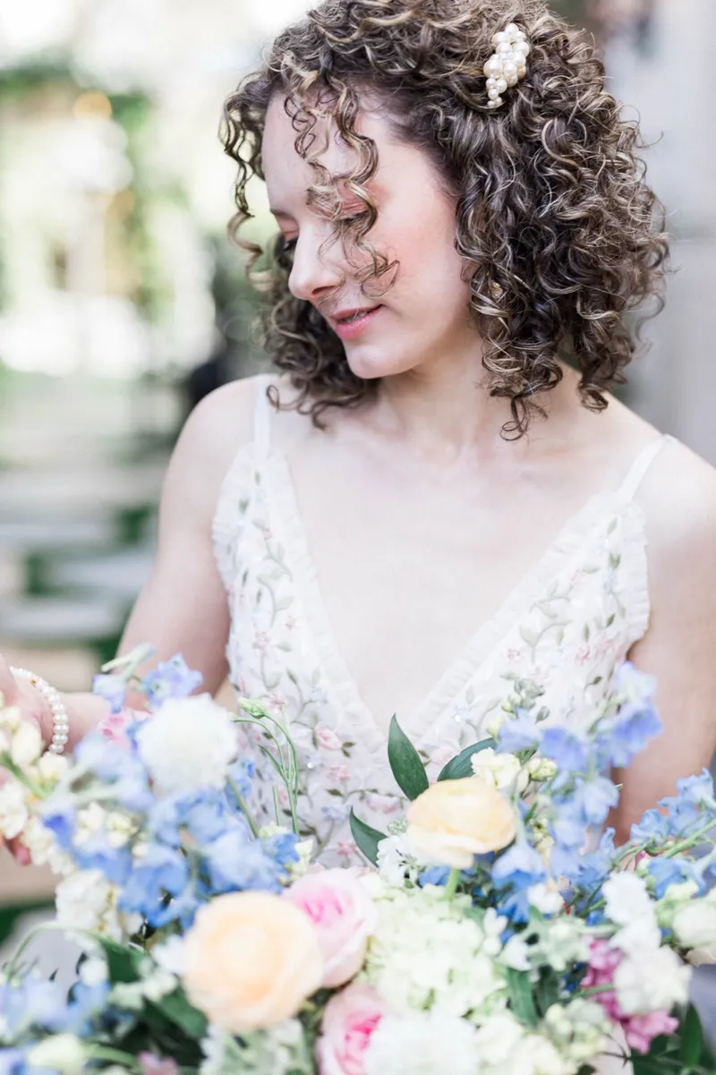 Close portrait of the bride smiling down at the blue and peach flowers, a pearl pin in her curls
