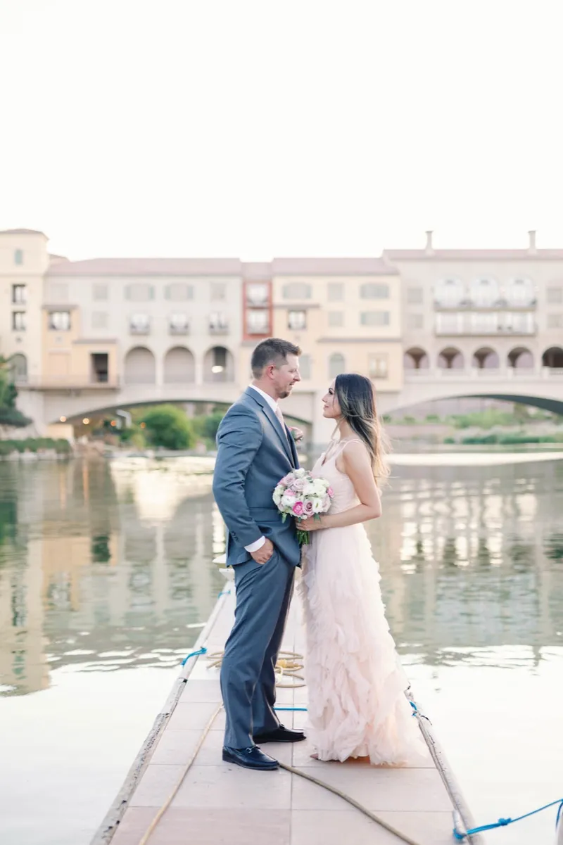 Couple standing together at the end of a dock with the village and the arched bridge across the water