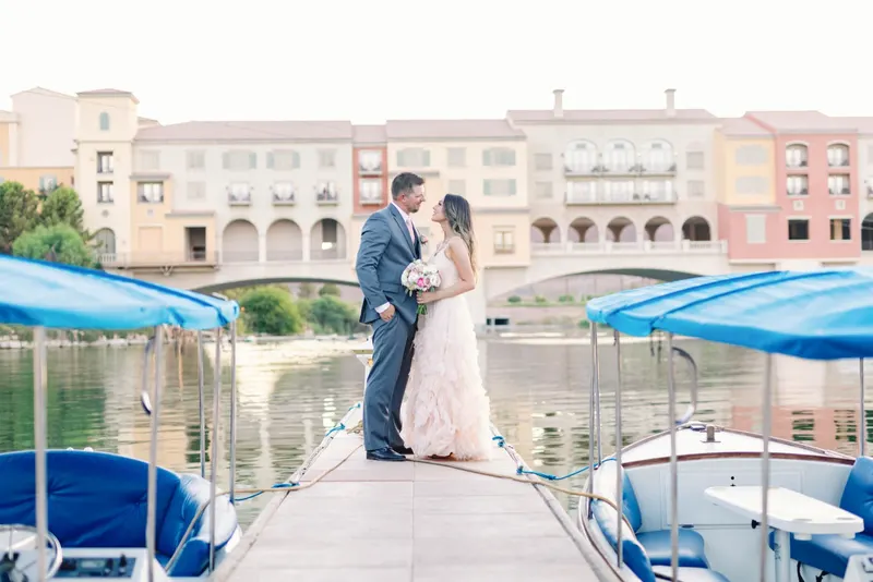 Bride in a blush ruffled gown and groom in a grey suit stand on a marina dock, village and stone bridge across the water