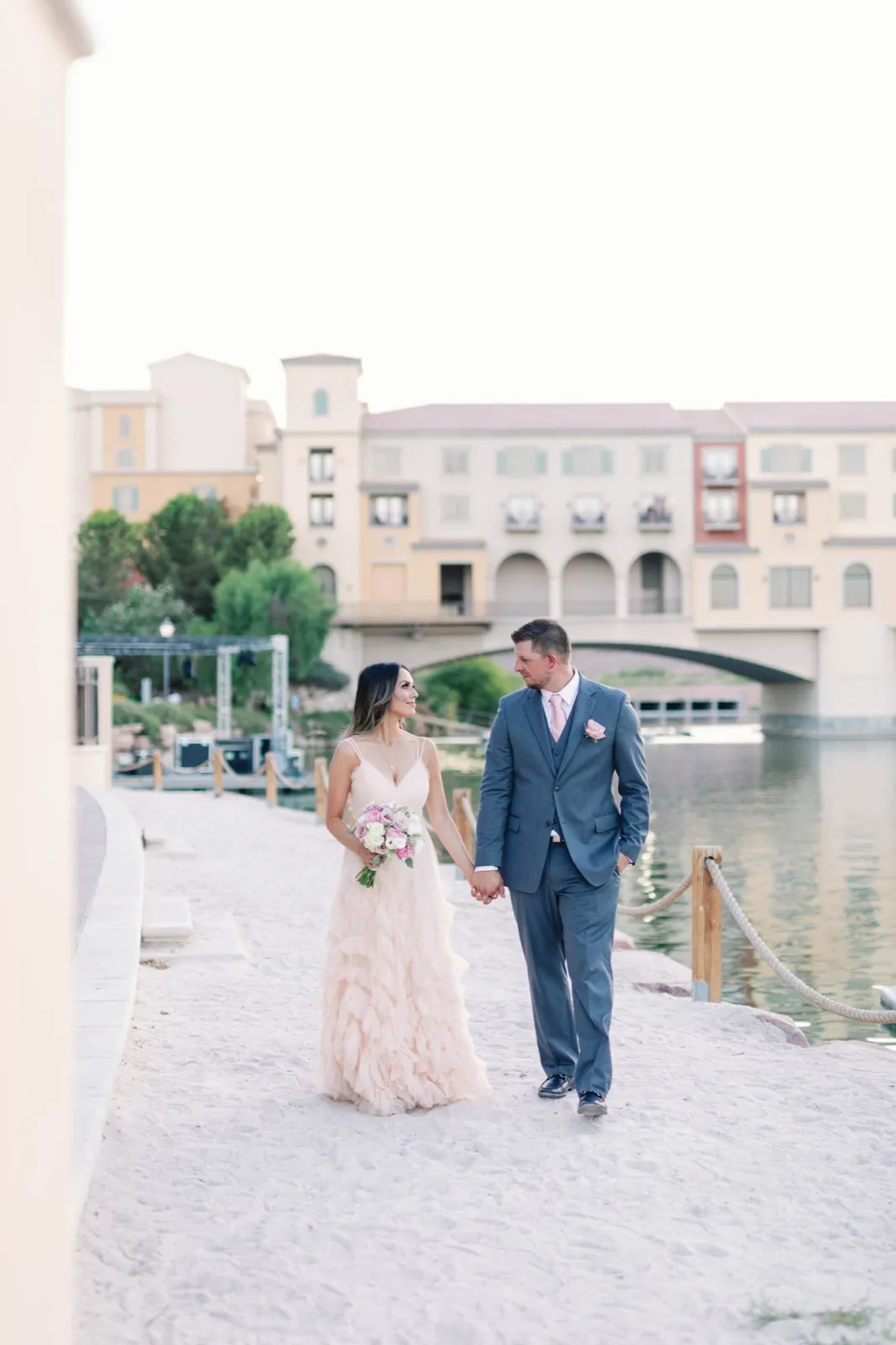 Bride in a blush gown and groom in grey walk a white sand shoreline, the village and stone bridge behind