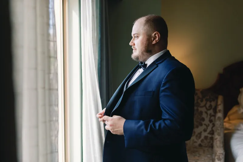 Groom buttoning his navy tuxedo jacket at a tall window with the curtain pulled back