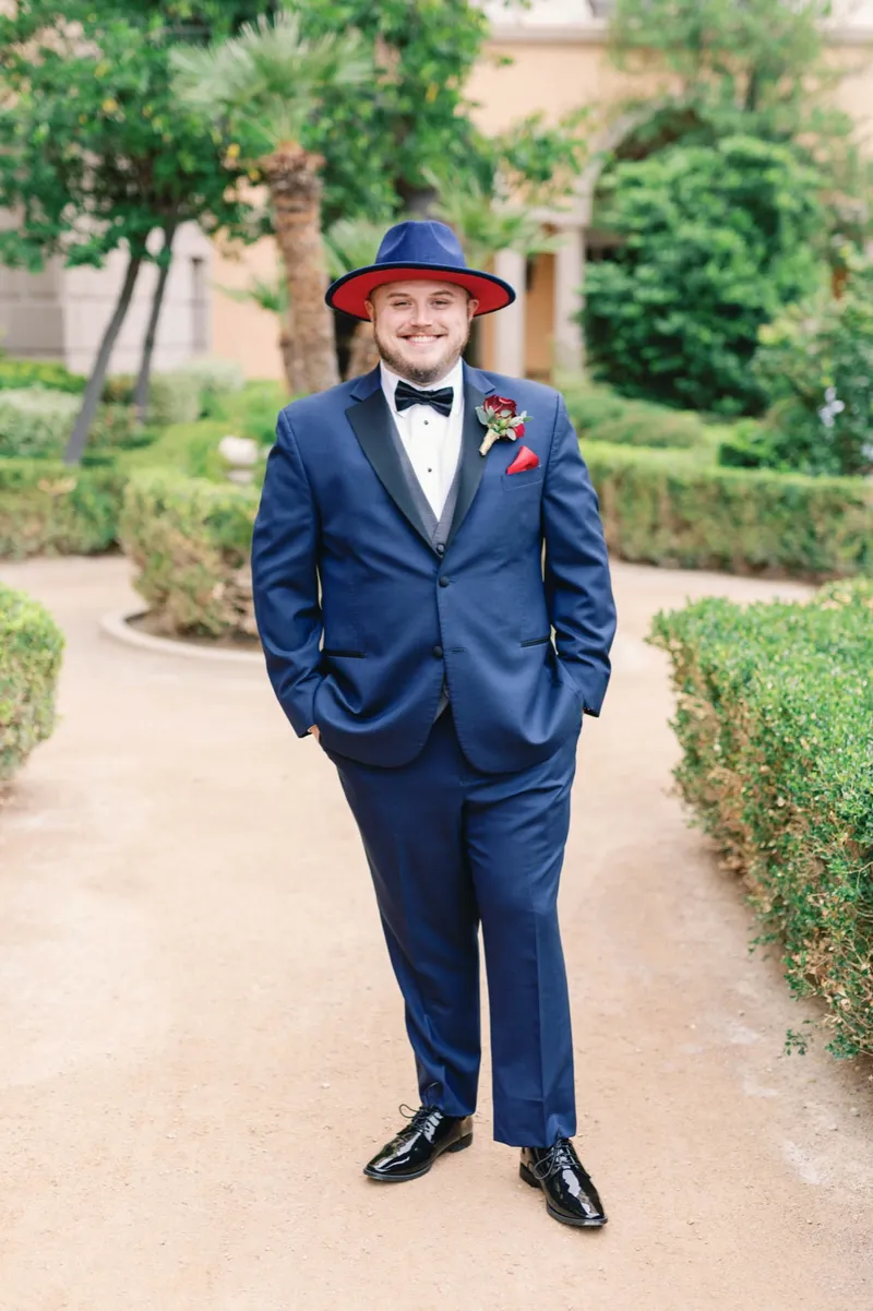 Groom standing on a garden path between clipped hedges in a navy tuxedo, a black bow tie and a navy hat with a red underbrim