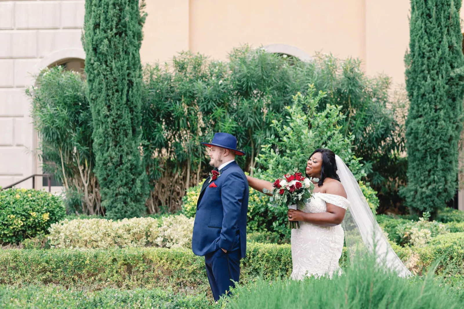Groom in a navy suit and hat waits with his back turned as his bride steps up behind him in a hedge garden