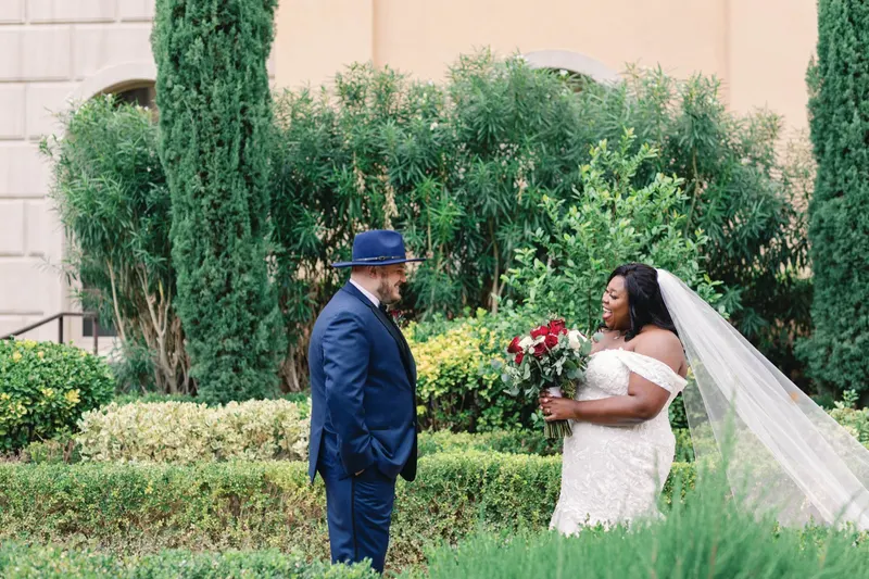 Bride laughing as she sees her groom for the first time in a hedge garden, him in a navy suit and hat