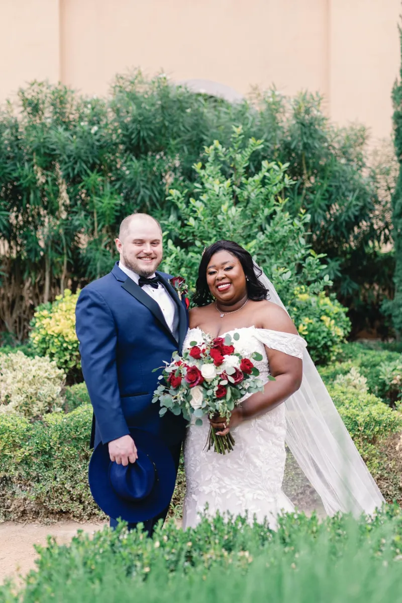 Bride and groom standing shoulder to shoulder in the garden, her bouquet in both hands and his hat in his