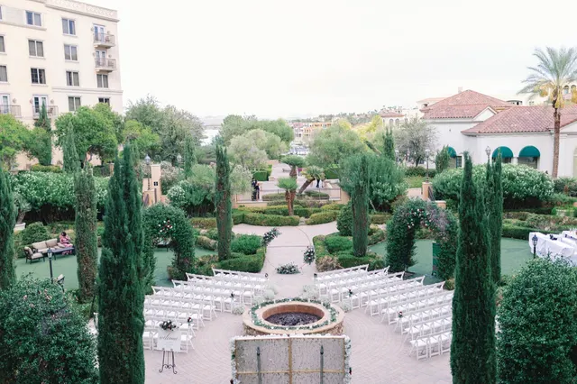 White chairs set in a fan around a round stone fire feature in a formal garden of cypress and clipped hedges, with the lake and the village rooftops in the distance