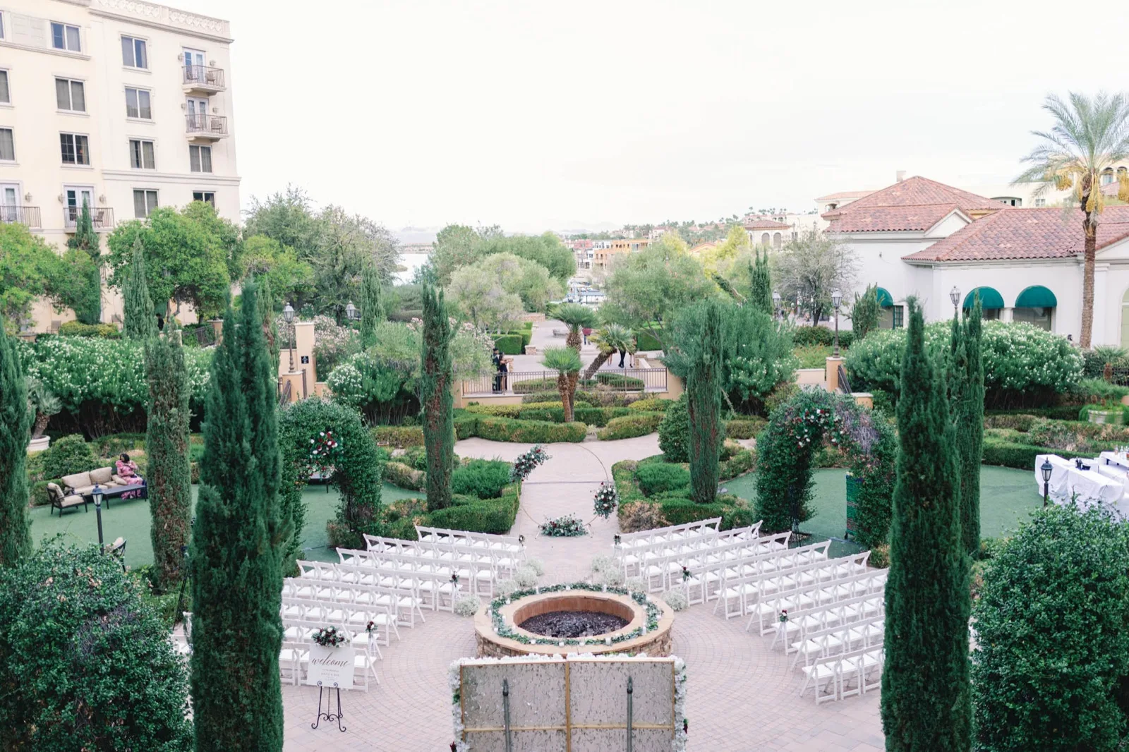 White chairs set in a fan around a round stone fire feature in a formal garden of cypress and clipped hedges, with the lake and the village rooftops in the distance