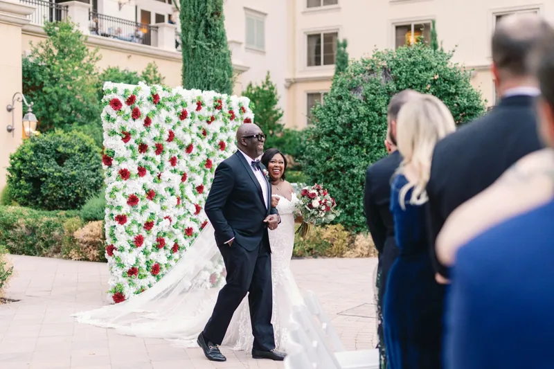Bride walking in on the arm of a man in a black tuxedo past a standing wall of white and red flowers
