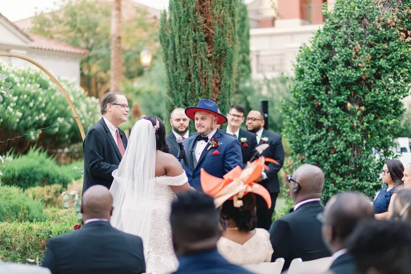Bride in an off shoulder lace gown facing her groom while their officiant reads from a book, groomsmen behind them
