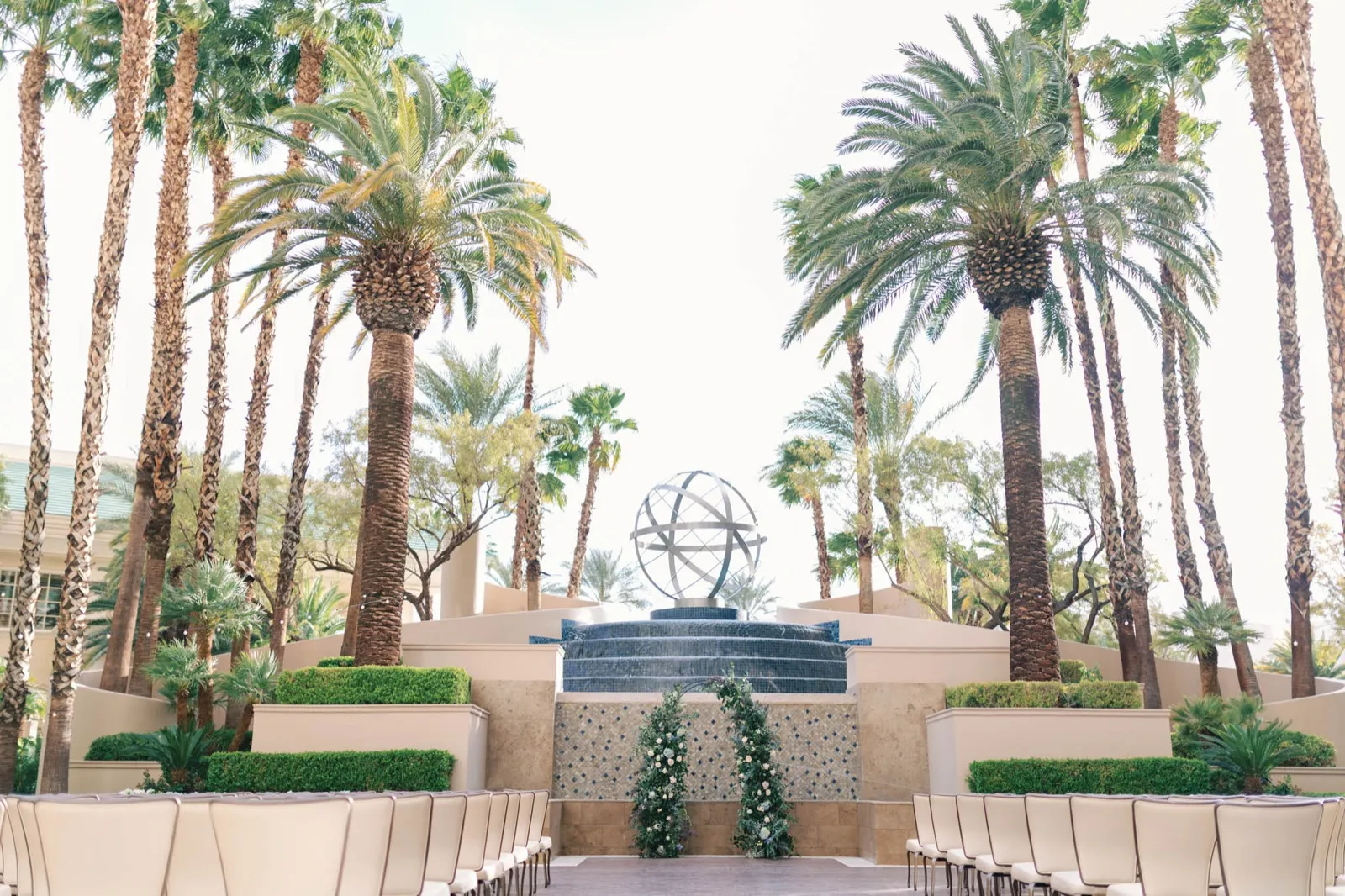 Rows of chairs facing a floral arch set in front of a tiered waterfall and sphere sculpture, ringed by tall palms