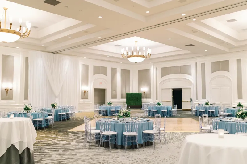 Ballroom set for dinner with round tables in dusty blue linens, clear chairs and an open dance floor under gold chandeliers
