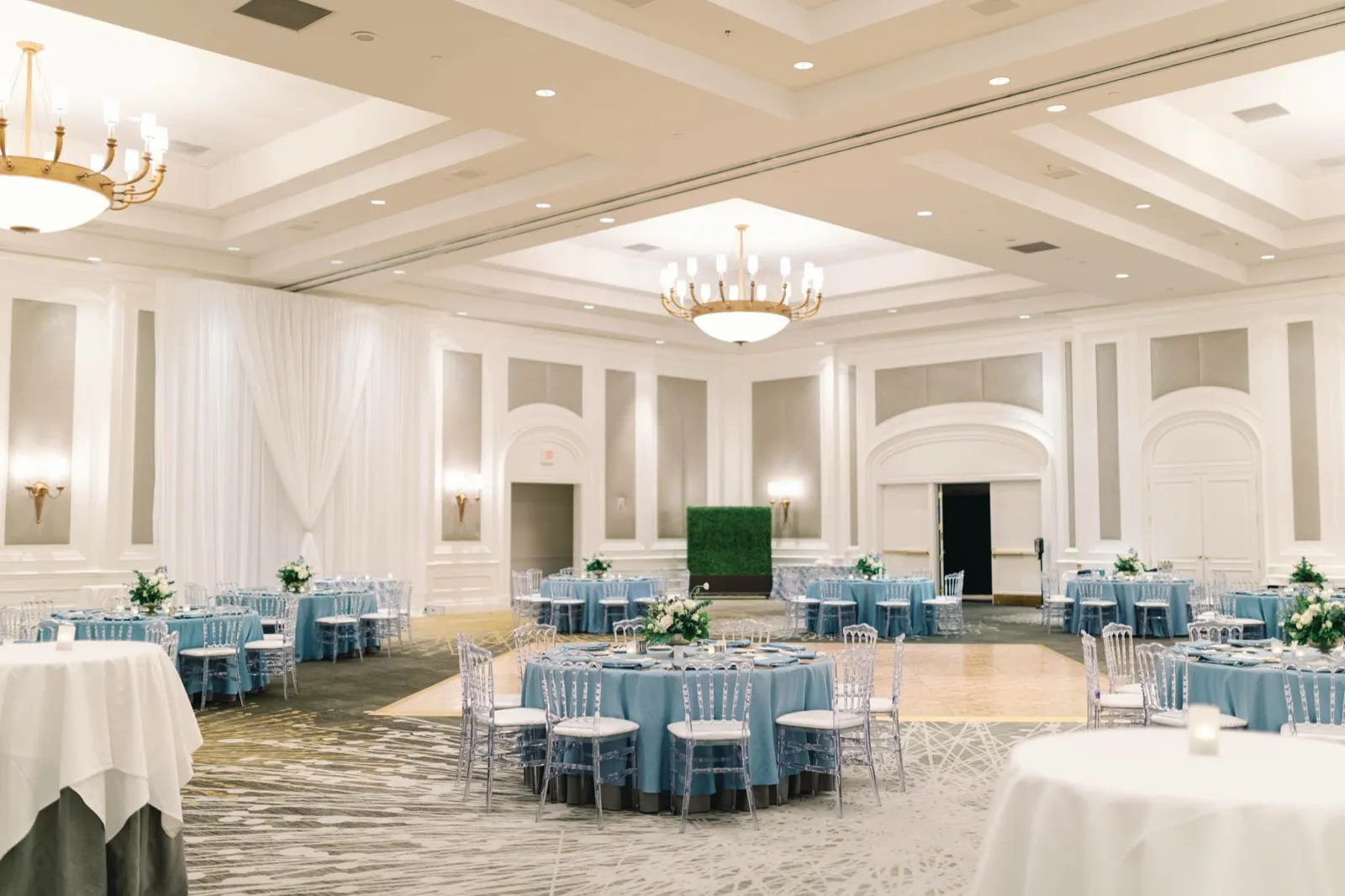 Hotel ballroom set for dinner: round tables in dusty blue linens, clear chairs and a dance floor under gold chandeliers