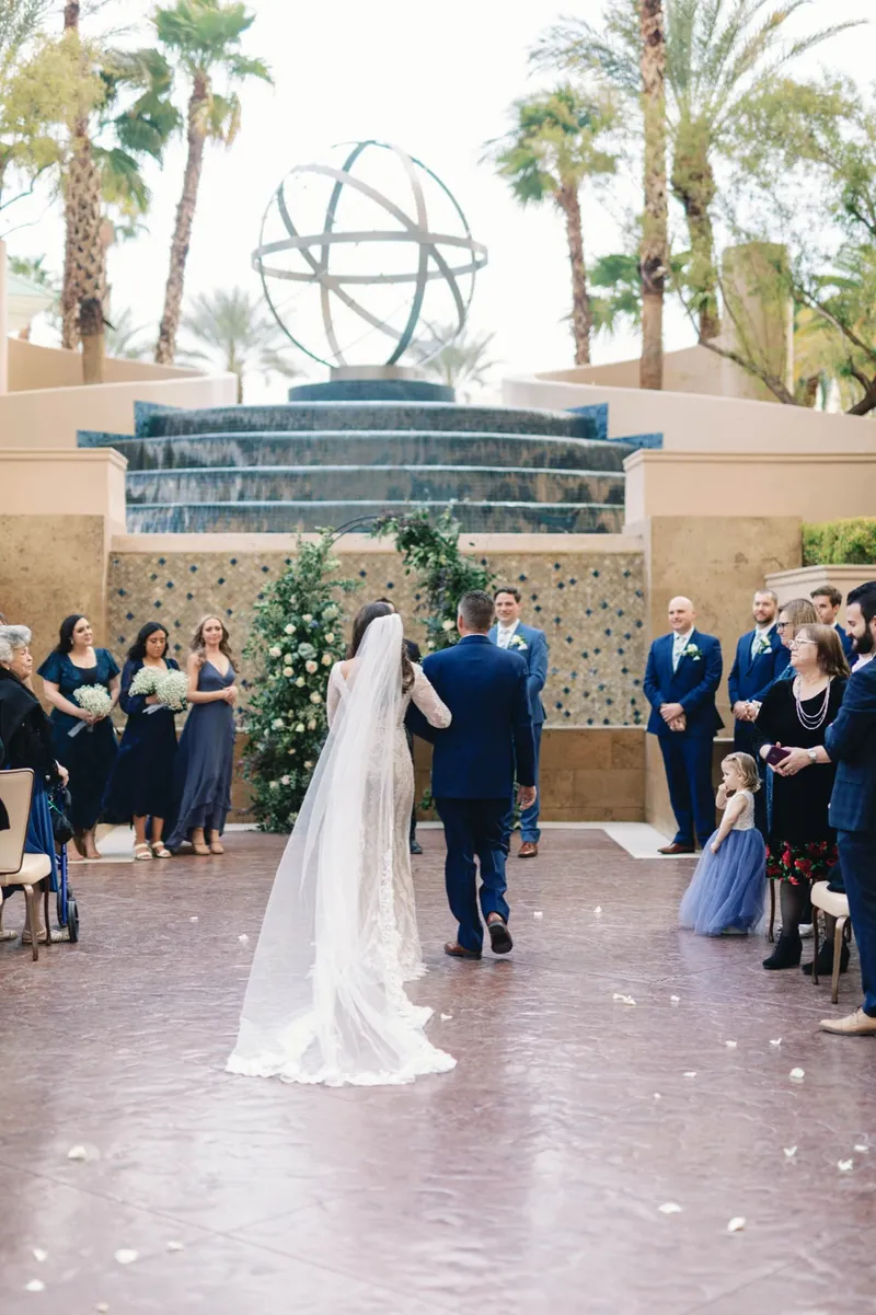 Bride walking up the aisle on her escort's arm toward the floral arch, bridesmaids in navy and a flower girl in periwinkle alongside