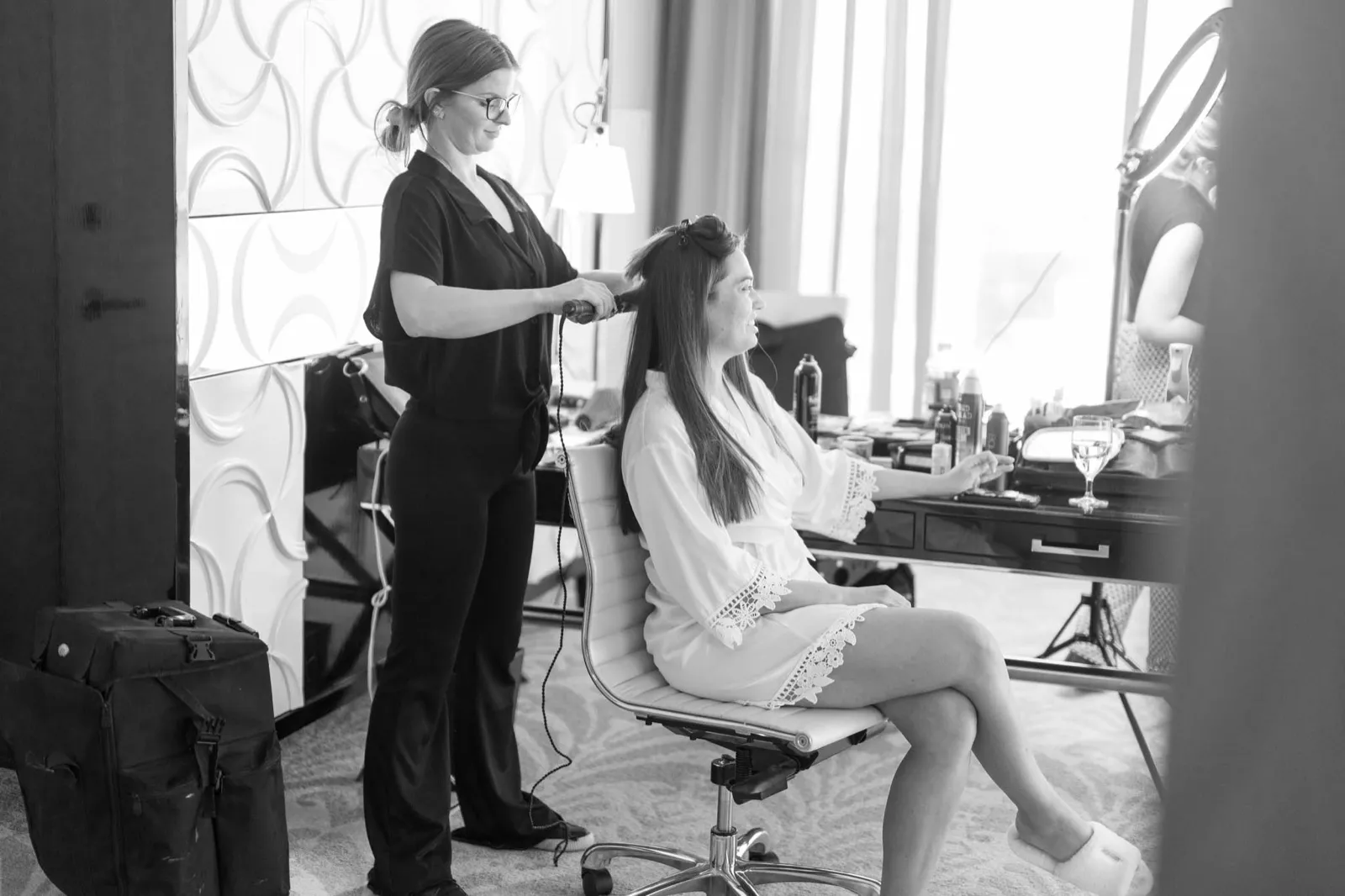 Black and white photo of a stylist curling the bride’s hair in a hotel suite beside a tall daylit window