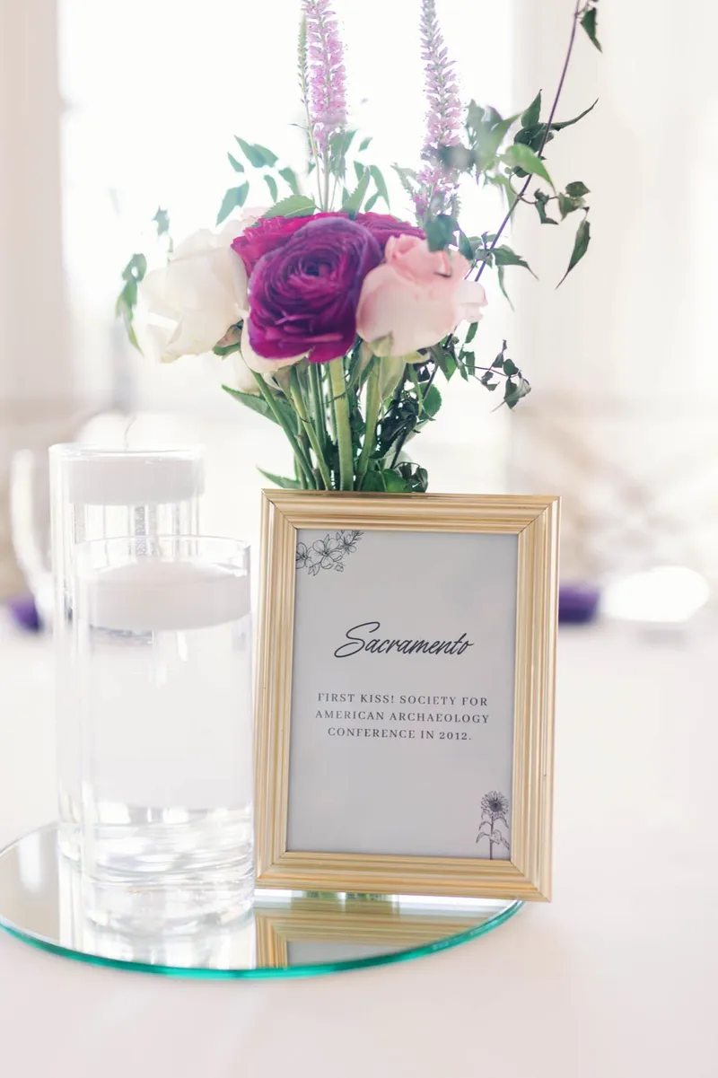 Framed card naming one of the reception tables after a city, standing beside a small arrangement of magenta and blush roses