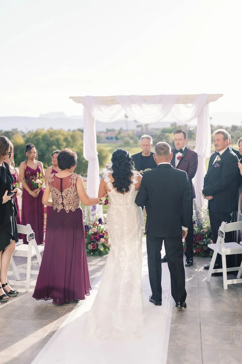 Bride walking down the aisle on the arms of her parents toward the draped arch