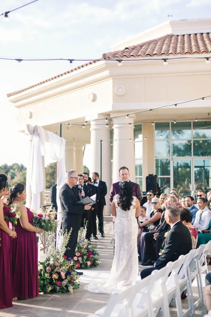 Terrace ceremony beside the clubhouse, the bride seen from behind facing her groom in a burgundy tuxedo while the officiant speaks