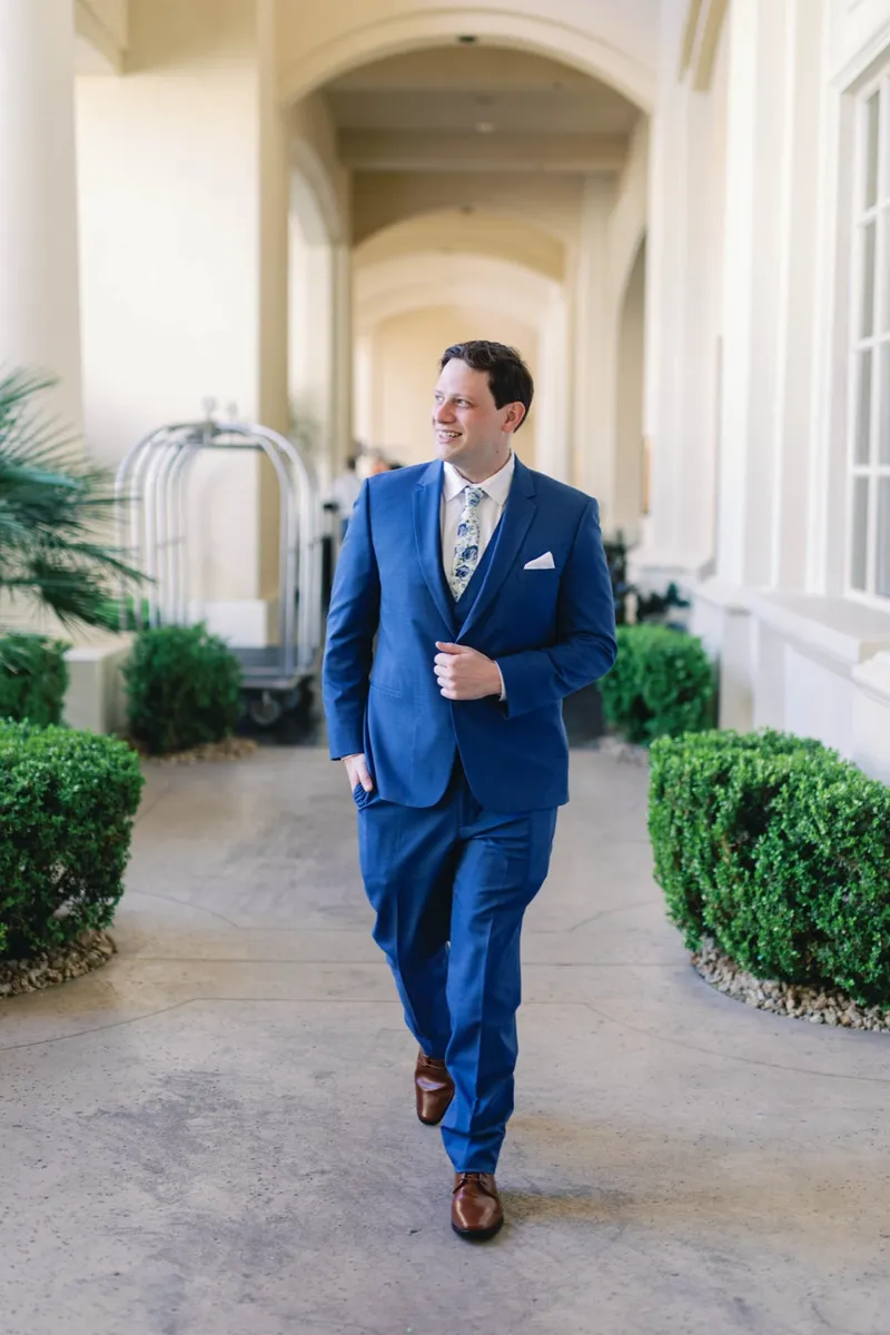 Groom in a blue suit walking toward the camera along the hedge lined walkway