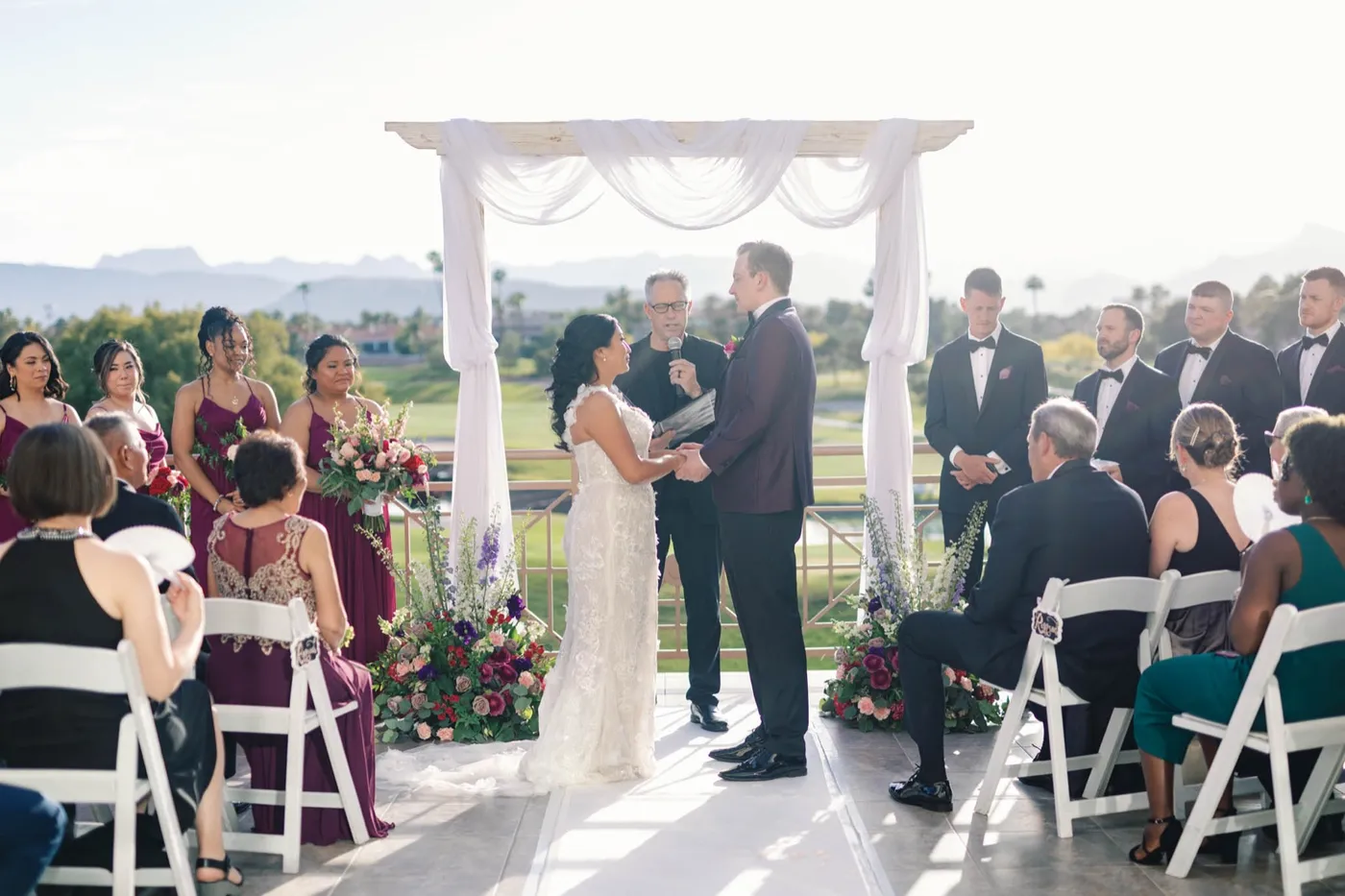 Couple exchange vows under a white draped arch on the patio, guests seated around, fairway and mountains behind