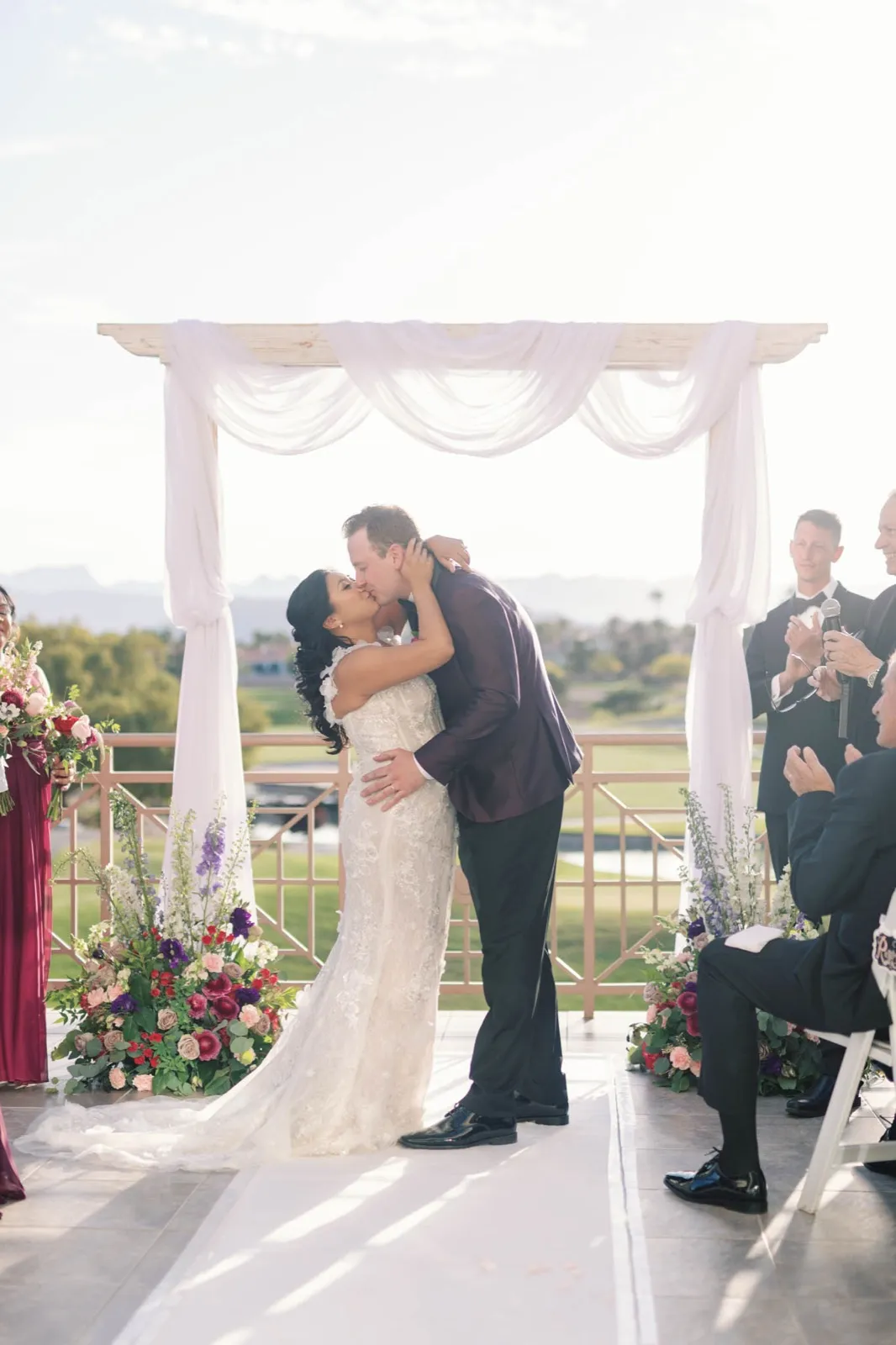 Couple share their first kiss under a white draped arch with the fairway and mountains behind them
