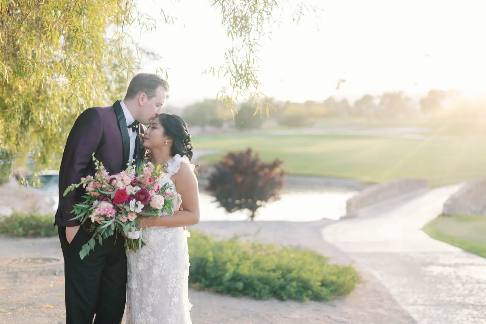 Groom kisses the bride’s forehead under a willow tree with the sunlit fairway and water behind them
