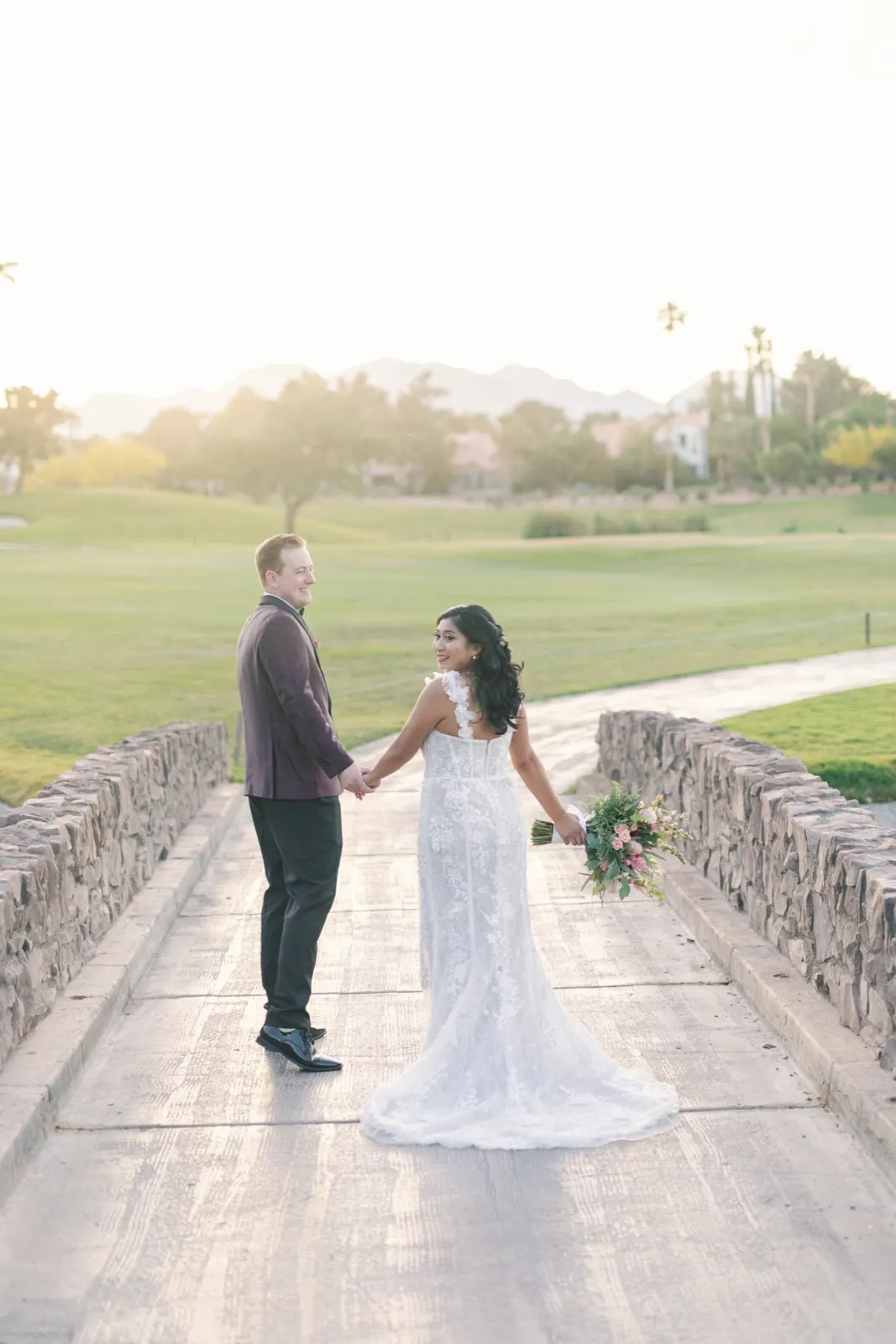 Bride looks back over her shoulder as the couple cross the stone bridge over the water at sunset