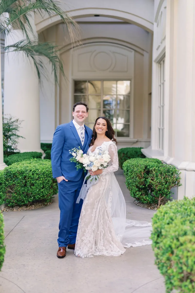 Bride and groom standing together between the clipped hedges outside the arched walkway