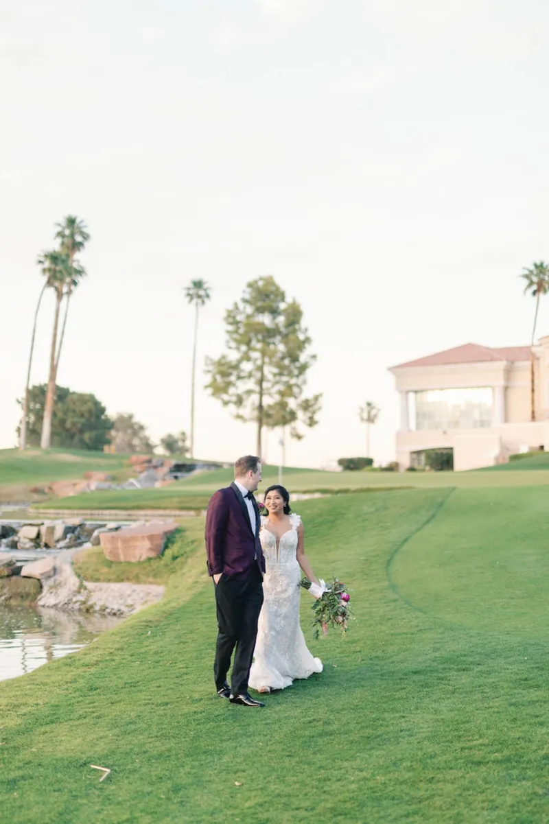 Couple walking the grass beside the water with palms and the clubhouse roof behind them