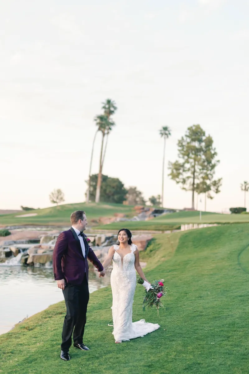 Couple standing on the slope beside the water hazard and its rock waterfall, the bride holding a magenta bouquet