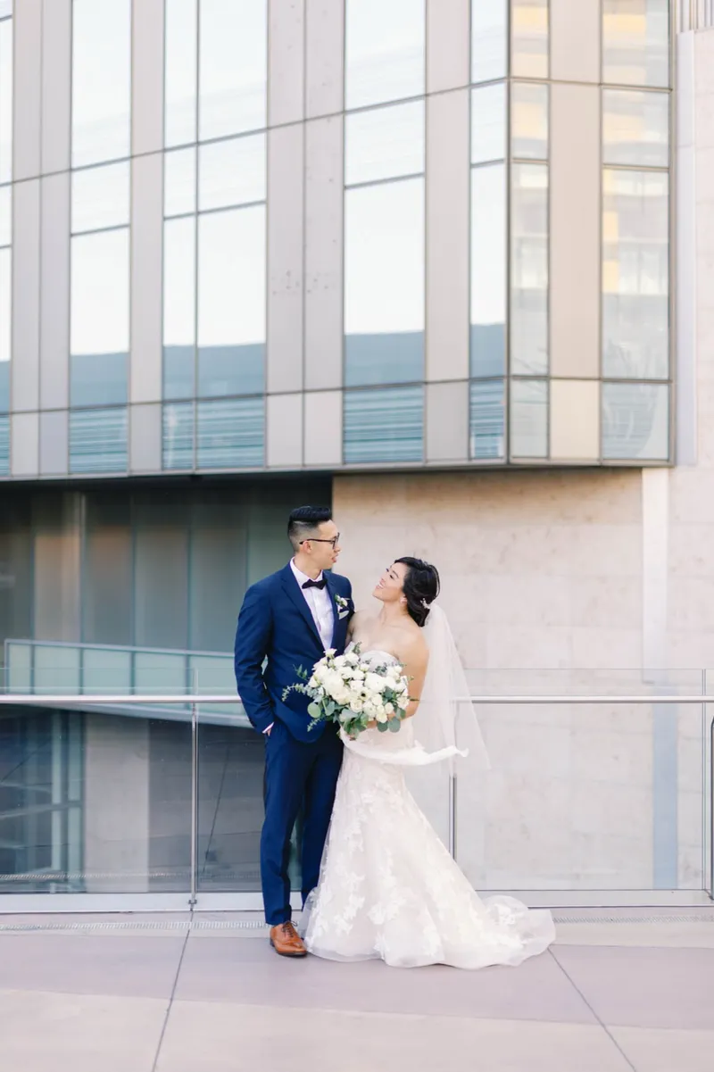 Bride and groom standing close together with the glass and pale stone walls of the resort rising behind them