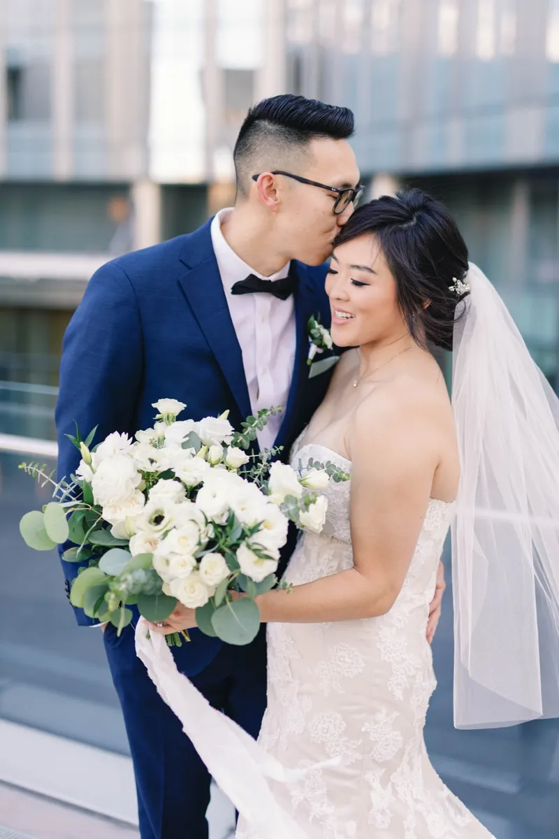 Groom kissing the bride's forehead as she holds her white and eucalyptus bouquet