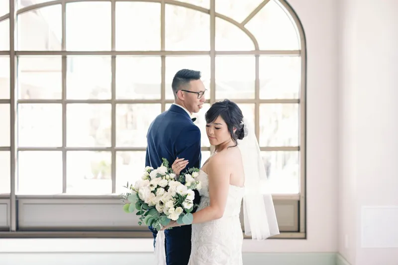 Bride and groom facing each other in front of a tall arched window with her bouquet between them