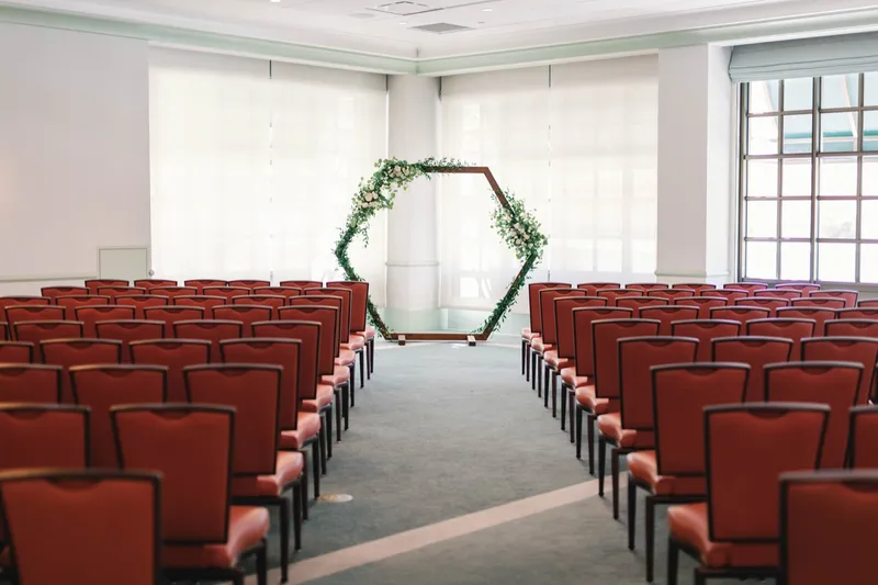 Ceremony room set with rows of rust red chairs facing a hexagon greenery arch, daylight coming through the tall windows