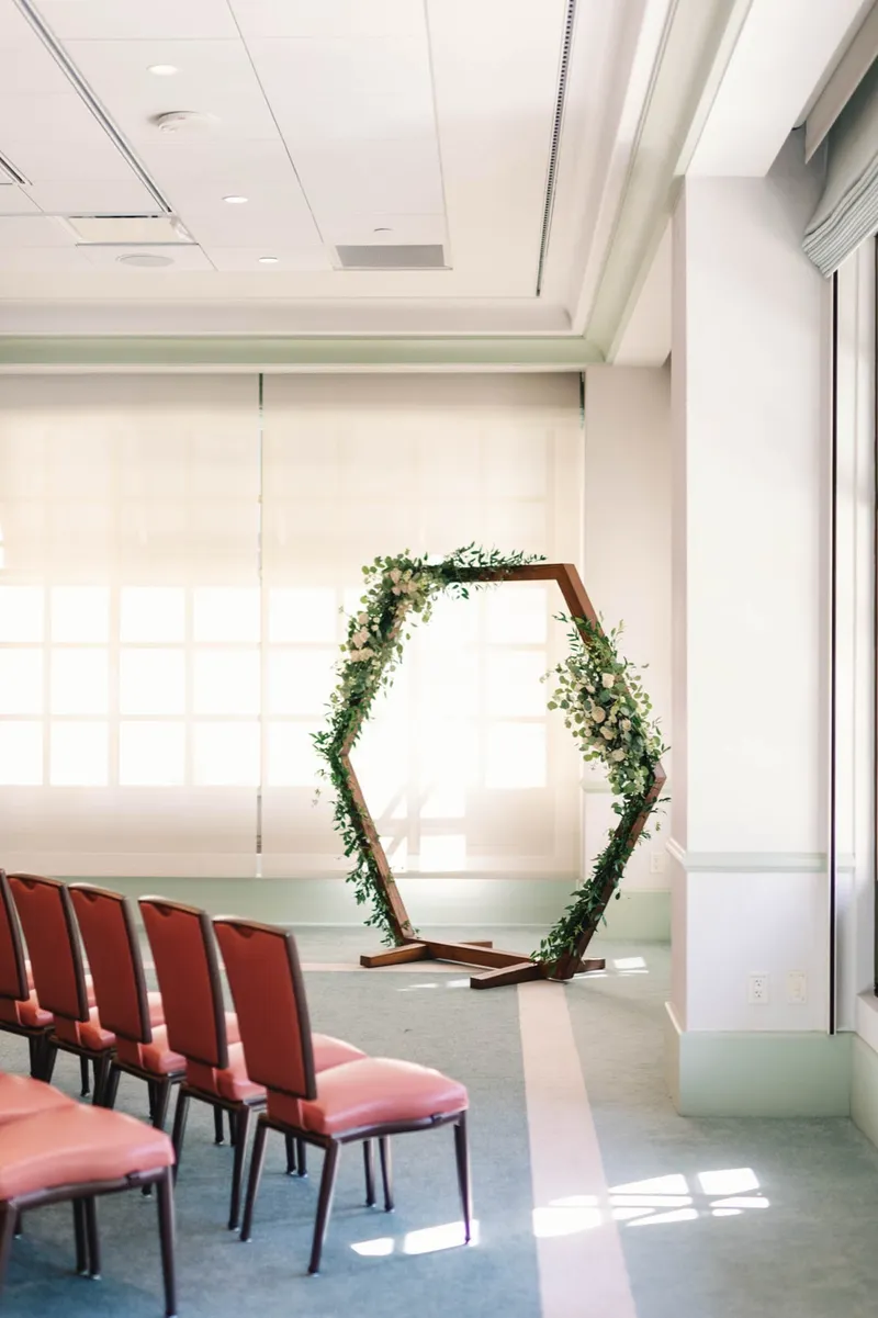 Hexagon arch wrapped in greenery and white blooms standing in the empty ceremony room before guests arrive