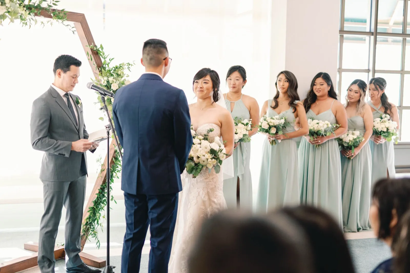 Officiant in a grey suit reads from a folded page at a microphone while a couple and six bridesmaids stand at a hexagon arch