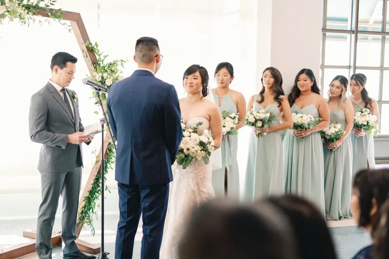 Bride and groom at the hexagon arch with the officiant beside them and bridesmaids in sage green