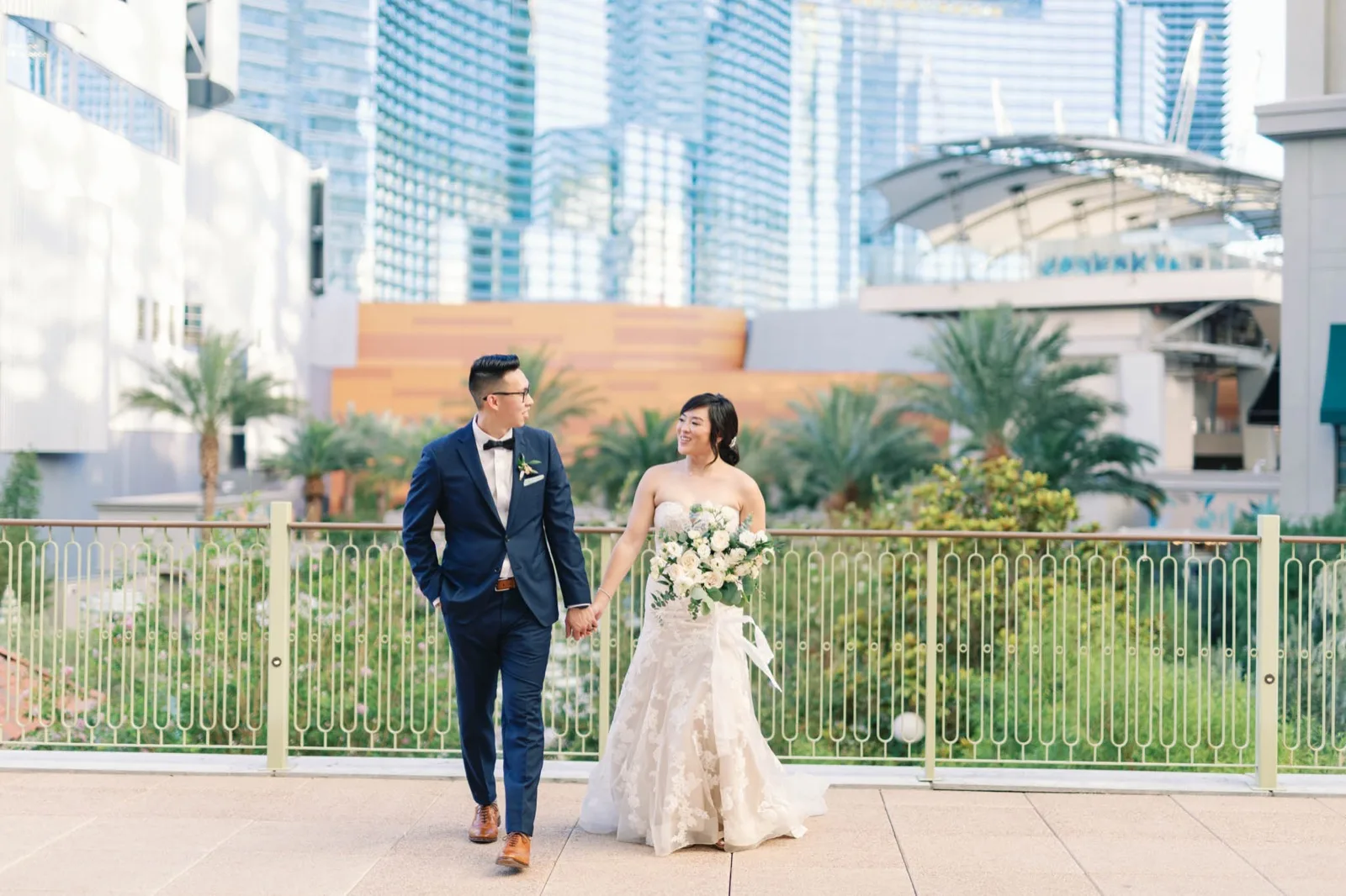 Bride and groom walking hand in hand along the terrace railing with palms below them and the glass towers of the Strip behind
