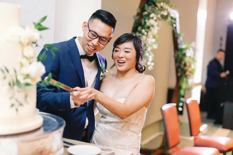 Bride and groom cutting their cake together in front of the greenery arch