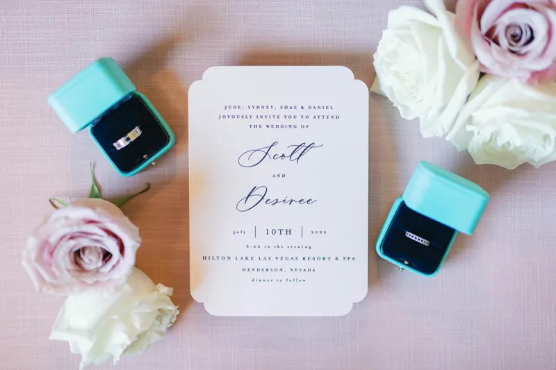 Wedding invitation laid on pale linen between two mint ring boxes holding a band and a diamond ring, with blush and white roses around it