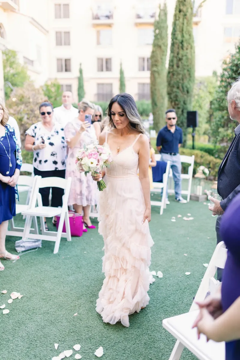 Bride walking herself down a grass aisle scattered with petals, her ruffled blush gown trailing behind her