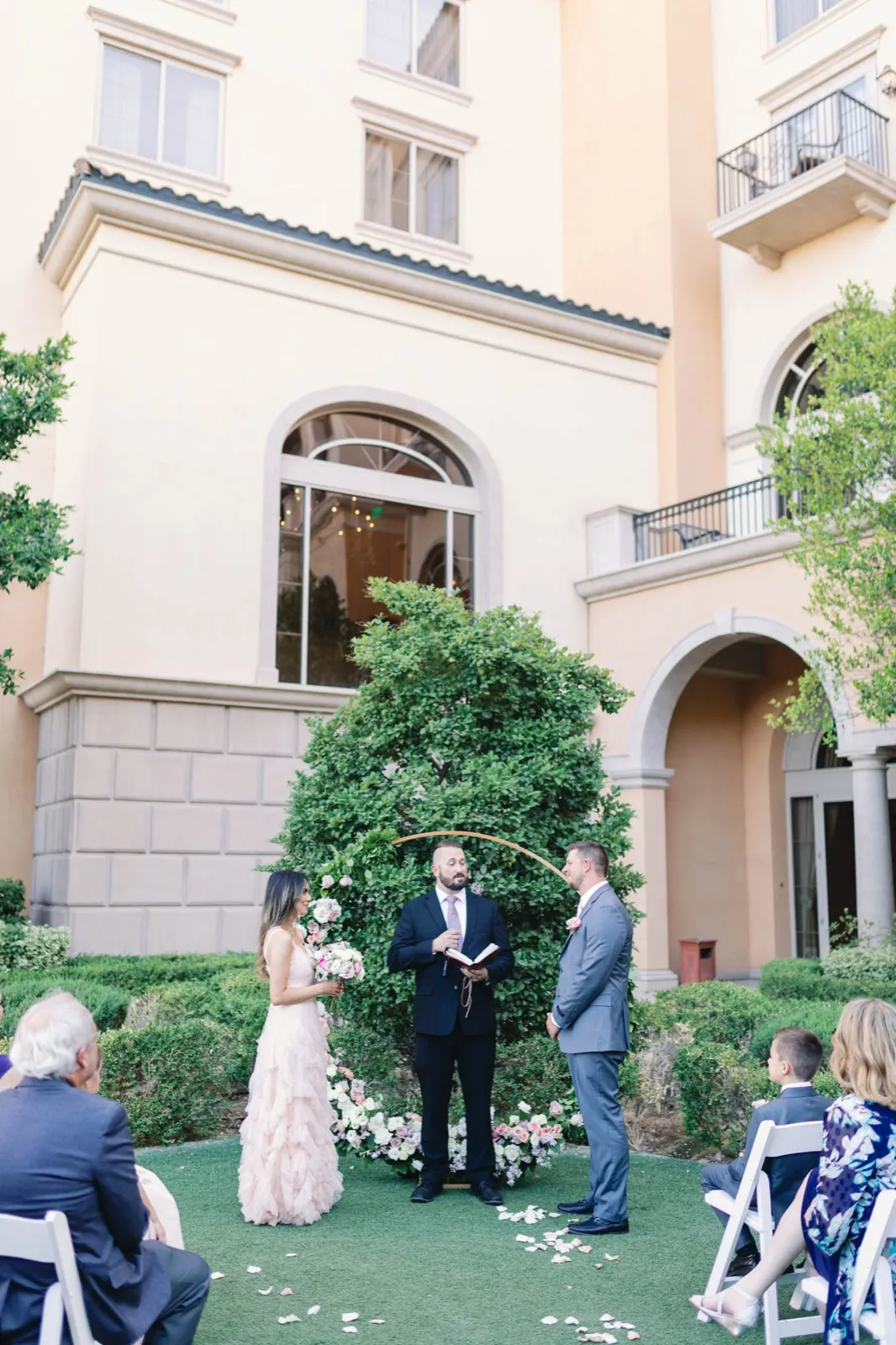 Bride, groom and officiant at a gold hoop arch of roses on a courtyard lawn below the resort’s arched facade