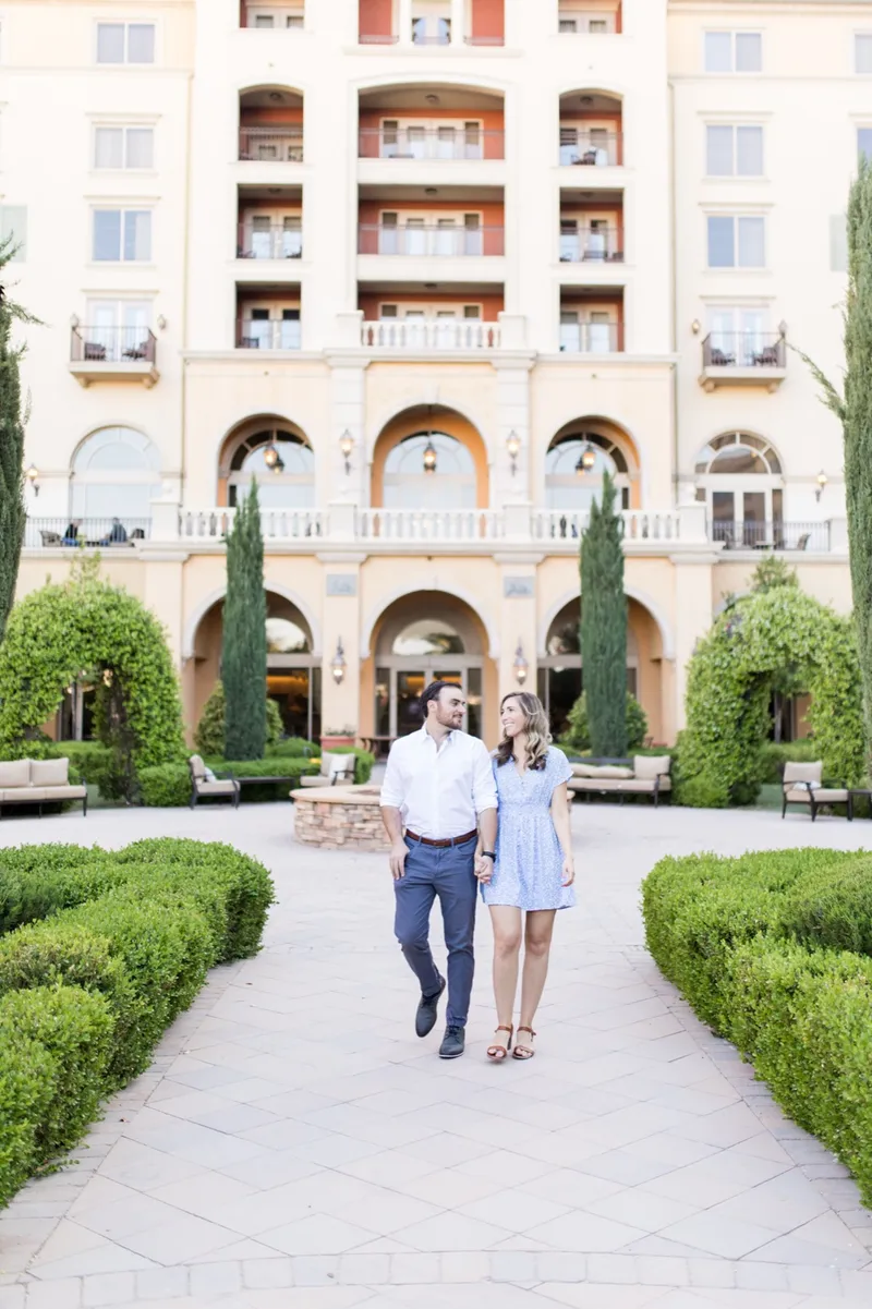 Couple walking toward the camera up a hedge lined courtyard path with a tall ochre resort building behind them