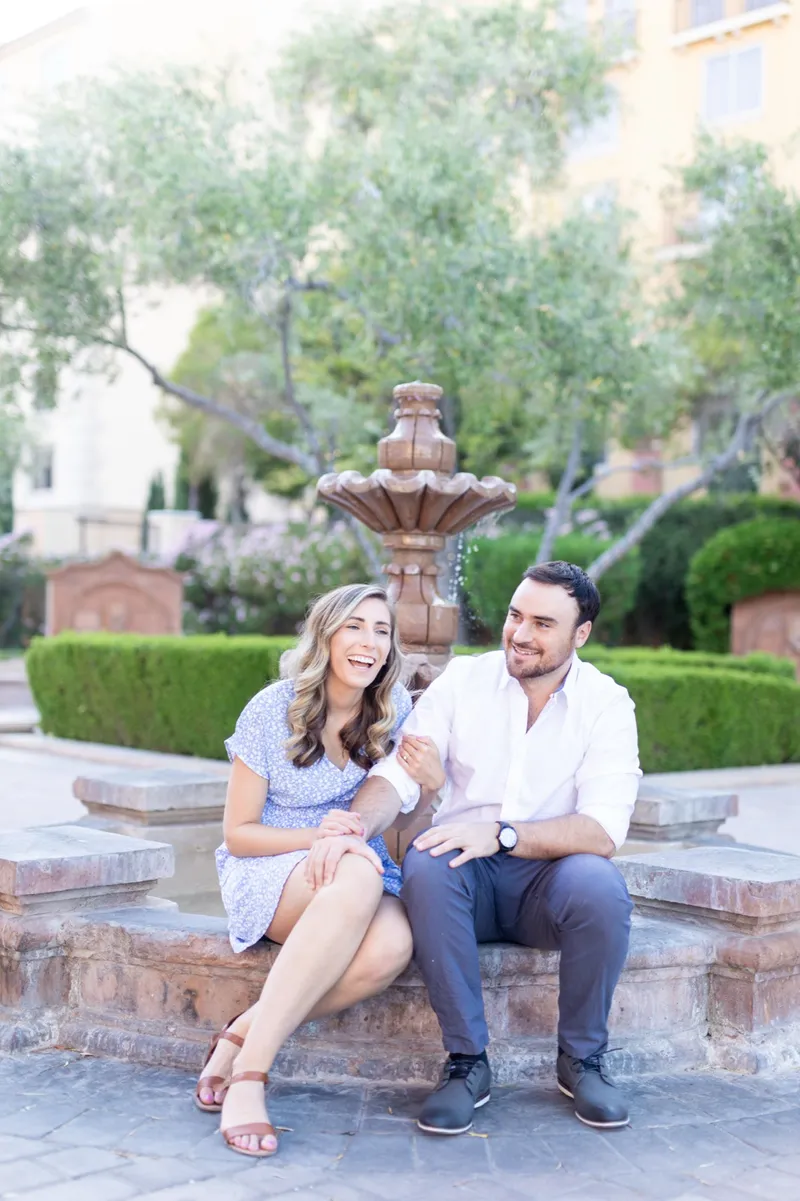 Couple sitting together on the stone rim of a courtyard fountain, both of them laughing